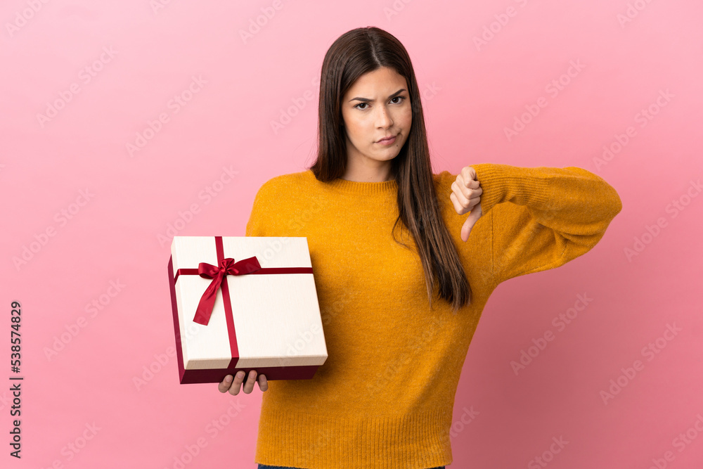 Teenager Brazilian girl holding a gift over isolated pink background showing thumb down with negative expression Teenager Brazilian girl holding a gift over isolated pink background showing thumb down with negative expression