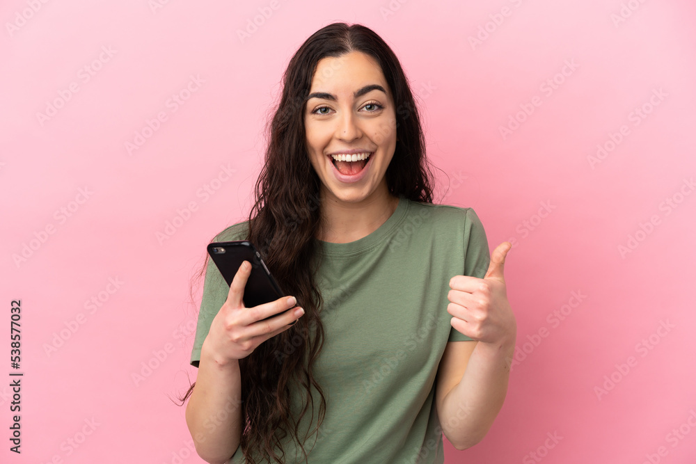 Young caucasian woman isolated on pink background using mobile phone while doing thumbs up Young caucasian woman isolated on pink background using mobile phone while doing thumbs up