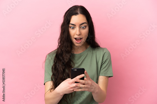 Young caucasian woman isolated on pink background looking at the camera while using the mobile with surprised expression