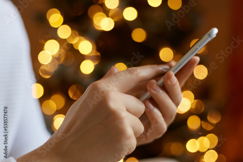 Unknown female hand holding mobile phone and typing, texting with friends, browsing Internet, anonymous woman posing with smart phone with blurred Christmas tree on background.