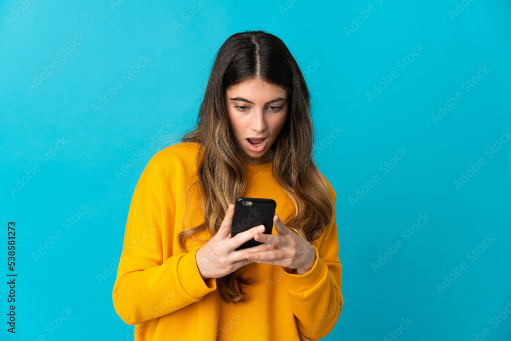 Young caucasian woman isolated on blue background looking at the camera while using the mobile with surprised expression Young caucasian woman isolated on blue background looking at the camera while using the mobile with surprised expression