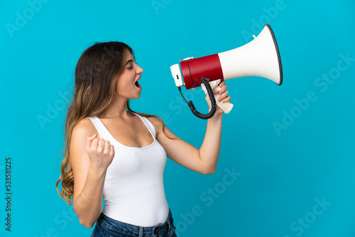 Young caucasian woman isolated on blue background shouting through a megaphone to announce something in lateral position