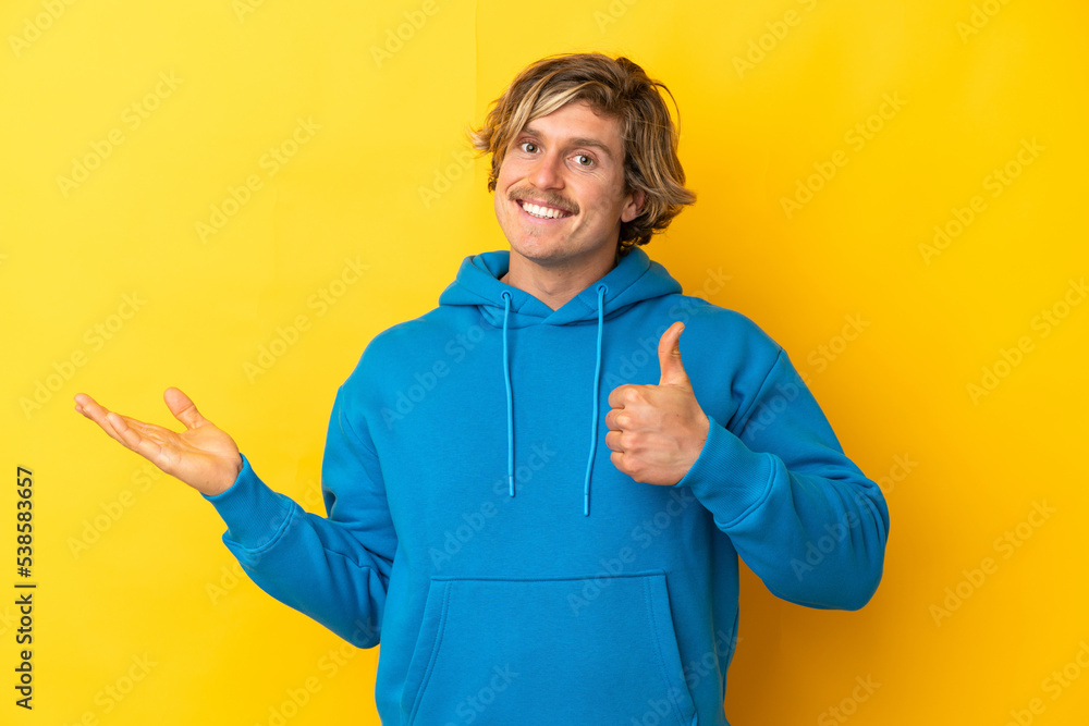Handsome blonde man isolated on yellow background holding copyspace imaginary on the palm to insert an ad and with thumbs up Handsome blonde man isolated on yellow background holding copyspace imaginary on the palm to insert an ad and with thumbs up