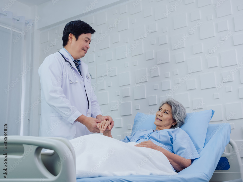 Fototapeta premium The kind Asian male doctor in white suit visits, talks and gives support, holding hand of happy elderly senior woman patient in light blue dress lying on bed in hospital room.