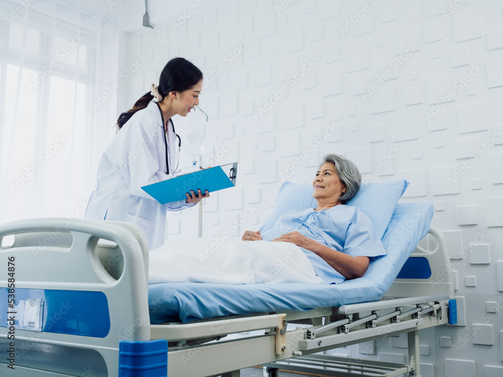 Obraz premium Young Asian female doctors visit senior woman patient in bed, explaining the symptoms and examination notes and medical healthcare documents on clipboard. Physician in white suit working in hospital.