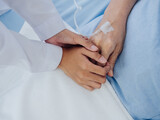 Close-up female doctor in white suit is holding on to elderly senior woman patient's hand in light blue dress with a saline hose lying on hospital bed, encouraging the patient concept.