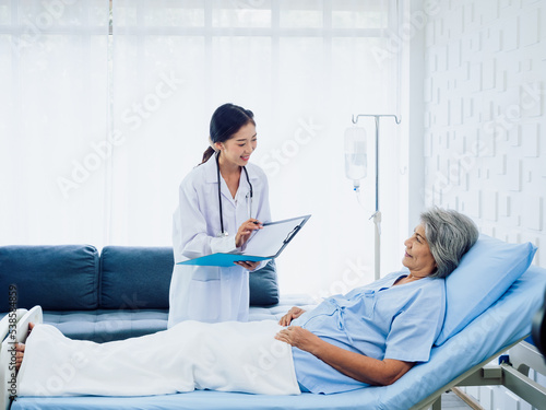 Young Asian female doctors visit senior woman patient in bed, explaining the symptoms and examination notes and medical healthcare documents on clipboard. Physician in white suit working in hospital.