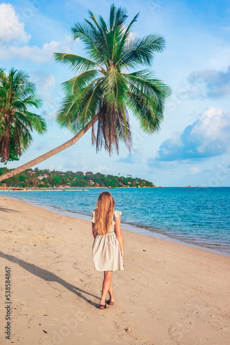 woman with long blond hair in a dress walks along a tropical seashore with palm trees. Travel and tourism