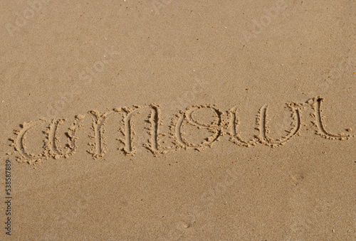 Close up on a message saying "amour" in French handwritten in the sand of a beach. Translation: "love. The texture of the sand is visible.
