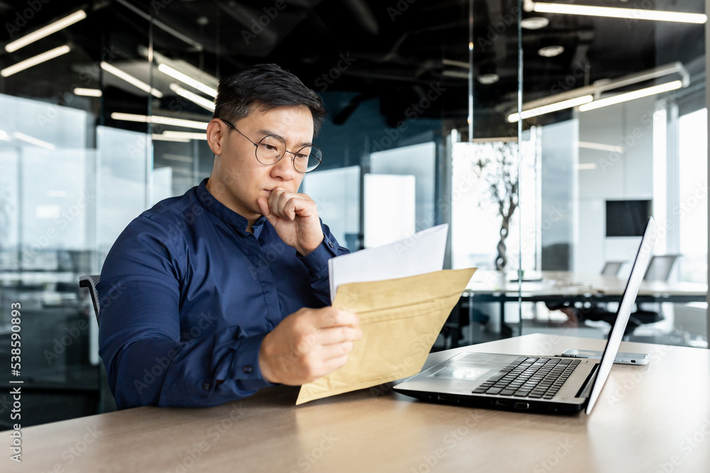 Thinking asian businessman working inside modern office building, man reading letter received from bank, boss thinking serious and concentrated working with laptop.