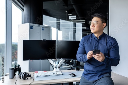 Fotografie Portrait of serious Asian architect businessman, man looking out window working in modern bright office inside using dual computer, businessman holding phone in hands