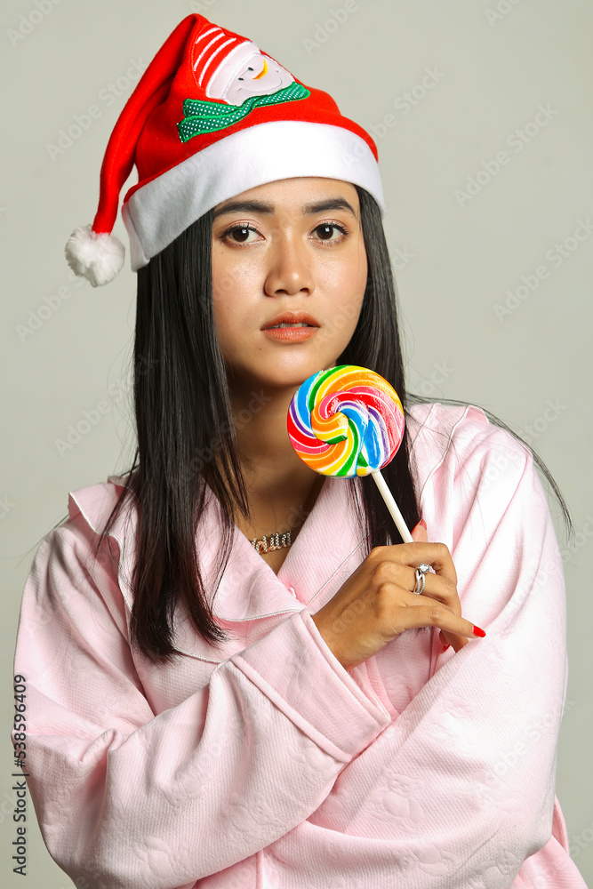 Fototapeta premium Portrait of beautiful young Asian woman wearing christmas hat and pink dress holding lollipop candy isolated on white background. christmas concept.