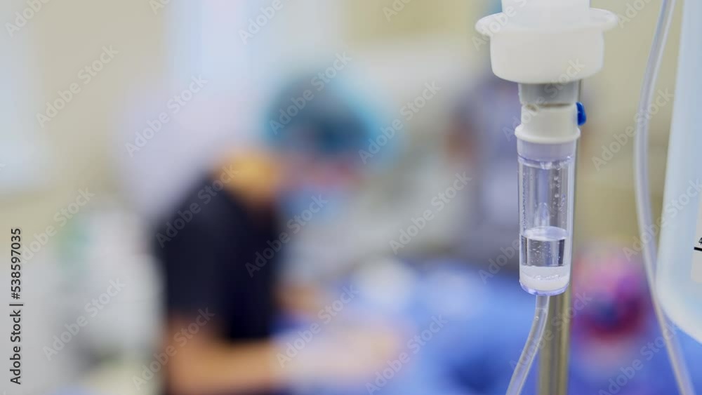 Apparatus for intravenous injections during surgeries. Medicine dripping in the drop counter in surgical room. Close up. Doctors work at backdrop in blur. Apparatus for intravenous injections during surgeries. Medicine dripping in the drop counter in surgical room. Close up. Doctors work at backdrop in blur.
