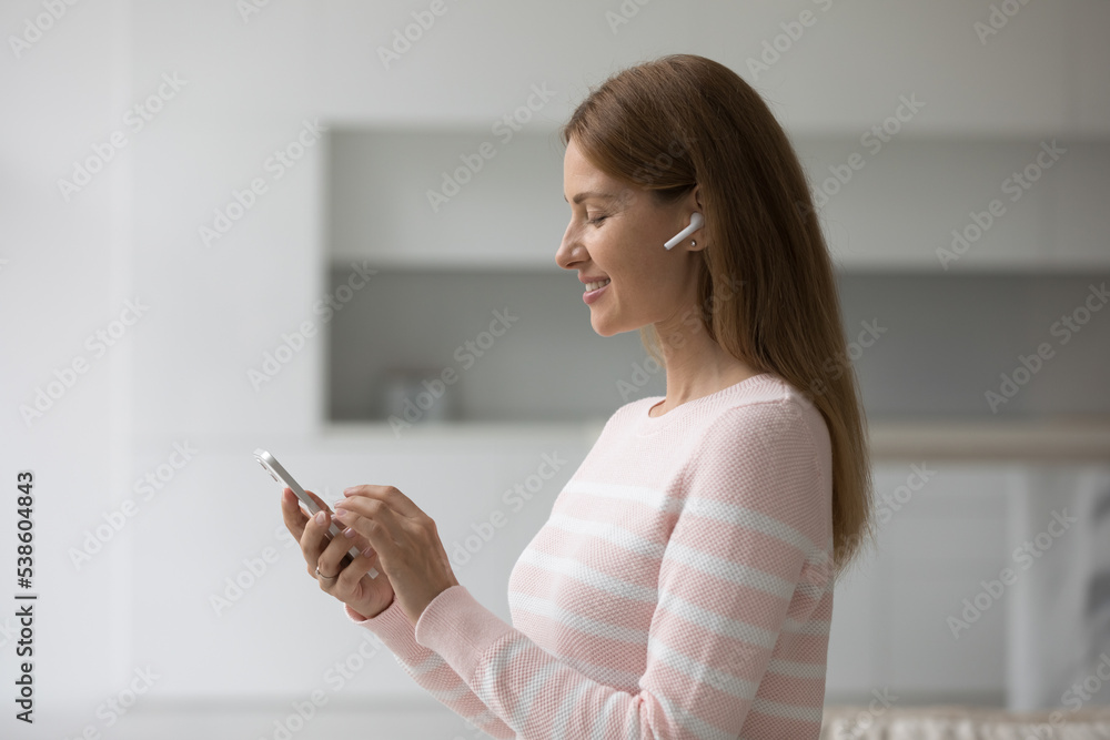 Attractive young woman standing in living room wear wireless earphones looking at smartphone screen smile enjoy favourite music, choose song, listen podcast spend leisure at home use modern technology Attractive young woman standing in living room wear wireless earphones looking at smartphone screen smile enjoy favourite music, choose song, listen podcast spend leisure at home use modern technology