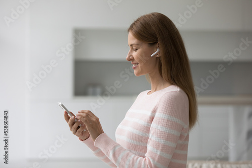 Attractive young woman standing in living room wear wireless earphones looking at smartphone screen smile enjoy favourite music, choose song, listen podcast spend leisure at home use modern technology