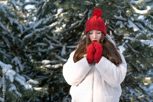 Beautiful young woman in knitted red hat and mittens blowing on her hands, snowy frorest background.Girl in winter park photo