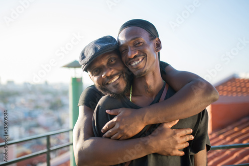 Close-up of happy African gay boyfriends looking at camera. Two bearded men in casual clothes standing together on roof top hugging taking photo on phone camera. LGBT peoples love, happiness concept