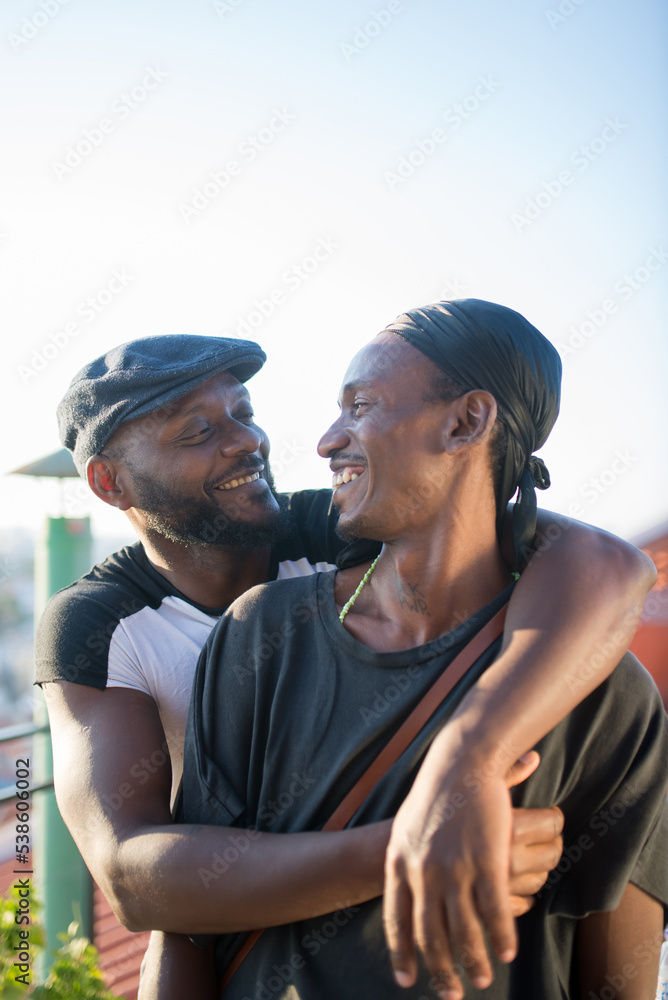 Portrait of romantic African gay couple hugging for photo. Two happy smiling men standing on roof top hugging looking at each other with pleasure. Same sex love and gay couples relations concept