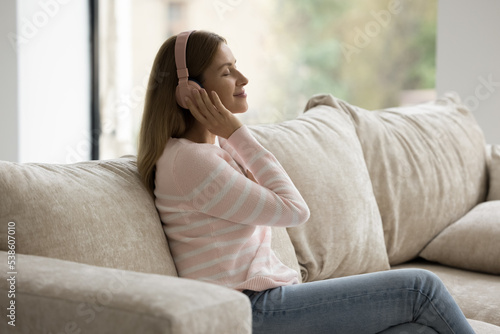 Young peaceful woman relaxing on sofa, enjoy track through wireless headphones, listen favourite song with eyes closed at home. Music lover spend stress-free leisure on weekend use modern tech, hobby