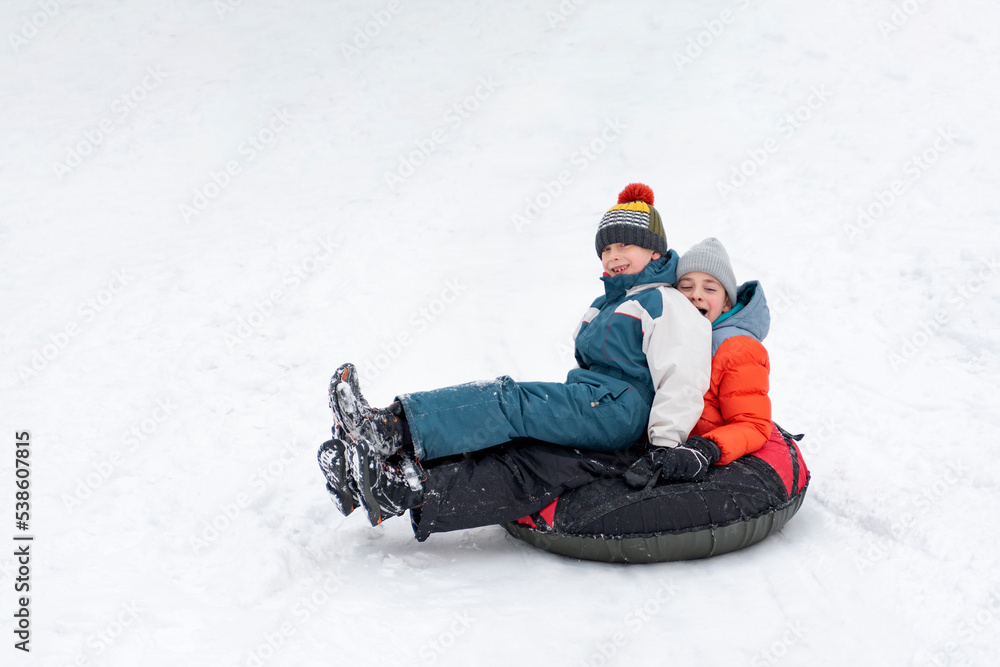 Children in winter jumpsuits and knitted hats slide down slope on inflatable sled. Boys have fun in winter outside. Children in winter jumpsuits and knitted hats slide down slope on inflatable sled. Boys have fun in winter outside.