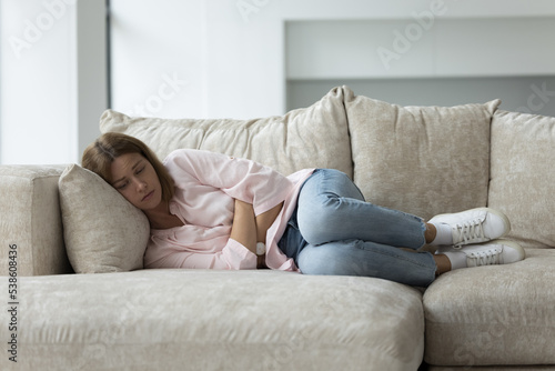 Sad unhappy young woman having physical malaise, suffers from painful feelings during period, lying on sofa hugs her belly resting at home. Psychological worries, divorce and break up, life failure