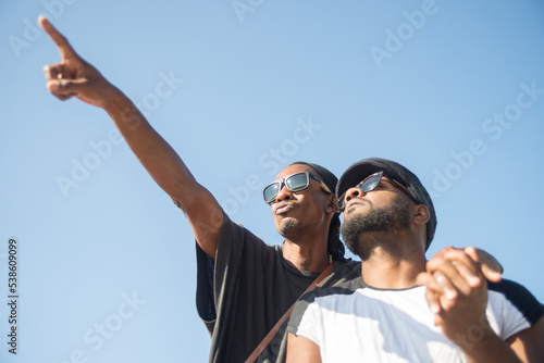 Portrait of happy African gay couple on blue sky background. Two bearded men standing together with hand in hand one man showing far away places with his finger. Same sex couples relationships concept