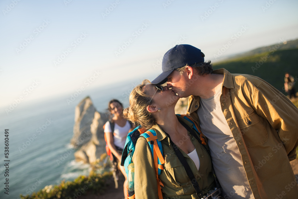 Happy mid adult couple kissing on top of mountain. Portrait of loving backpackers with children in background. Active family and relationships concept Happy mid adult couple kissing on top of mountain. Portrait of loving backpackers with children in background. Active family and relationships concept