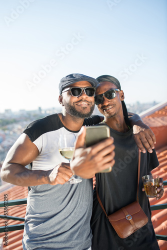 Portrait of African gay couple with smart phone on roof top. Two smiling bearded men in T-shirts standing close on balcony hugging and taking selfie. LGBT peoples love and happiness concept