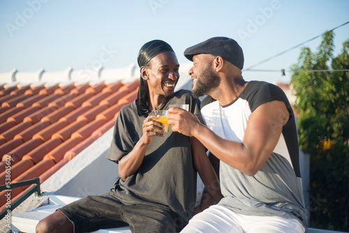 Portrait of African gay couple expressing love. Two bearded men dating on roof top sitting close smiling holding glasses of juice looking at each other with tender. LGBT couples love and life concept
