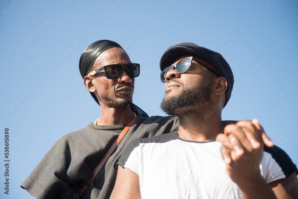Close-up of two happy African gay men dating. Handsome bearded men in sunglasses standing together holding hands and looking at each other on blue sky background. Concept of love care and LGBT rights Close-up of two happy African gay men dating. Handsome bearded men in sunglasses standing together holding hands and looking at each other on blue sky background. Concept of love care and LGBT rights