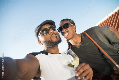 Close-up of two African gay men making faces on camera. Two men in sunglasses standing close on roof top with glasses of drinks and taking selfie. Same sex love, LGBT couples rights concept. .