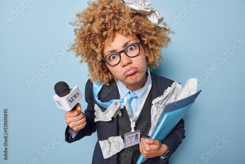 Journalism and breaking news concept. Serious displeased curly haired successful woman correspondent holds microphone folders and newspapers isolated over blue background going to take interview