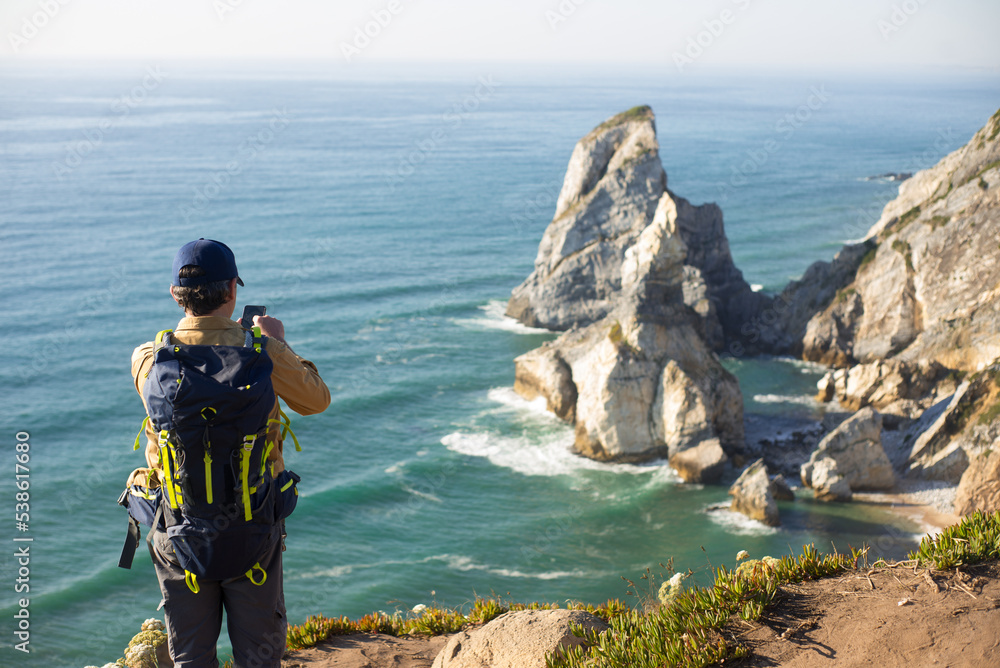 Rear view of backpacker taking photo of seascape. Mid adult man with backpack standing at precipice with smartphone enjoying view. Hiking weekend concept