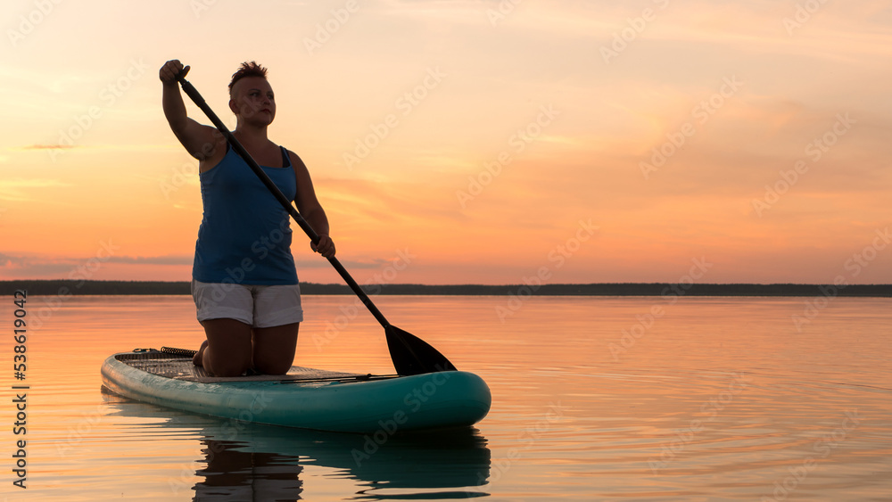 A woman with a Mohawk in shorts on her knees on a SUP board with an oar against the backdrop of a bright sunset sky swims in the lake in the evening.