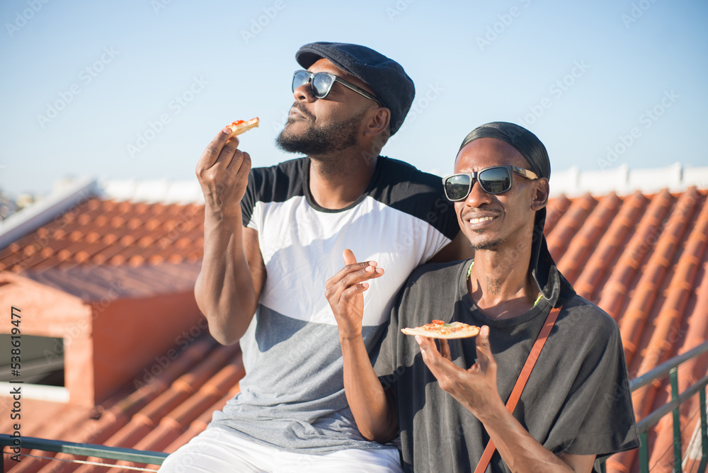 Portrait of happy African American gay men posing with pizza. Two boyfriends in sunglasses spending time on roof top eating pizza and enjoying summer weather. LGBT couples life and equality concept