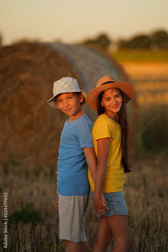 A boy and a girl stand with their backs to each other in bright summer clothes in the field and look smiling into the camera, in the background a haystack and a field. A boy and a girl stand with their backs to each other in bright summer clothes in the field and look smiling into the camera, in the background a haystack and a field.