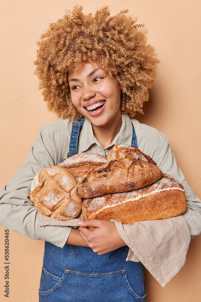 Vertical shot of positive curly haired woman carries loaves of bread on towel happy to bake fresh organic food wears shirt and denim apron isolated over beige background. Bakery shop worker. Vertical shot of positive curly haired woman carries loaves of bread on towel happy to bake fresh organic food wears shirt and denim apron isolated over beige background. Bakery shop worker.