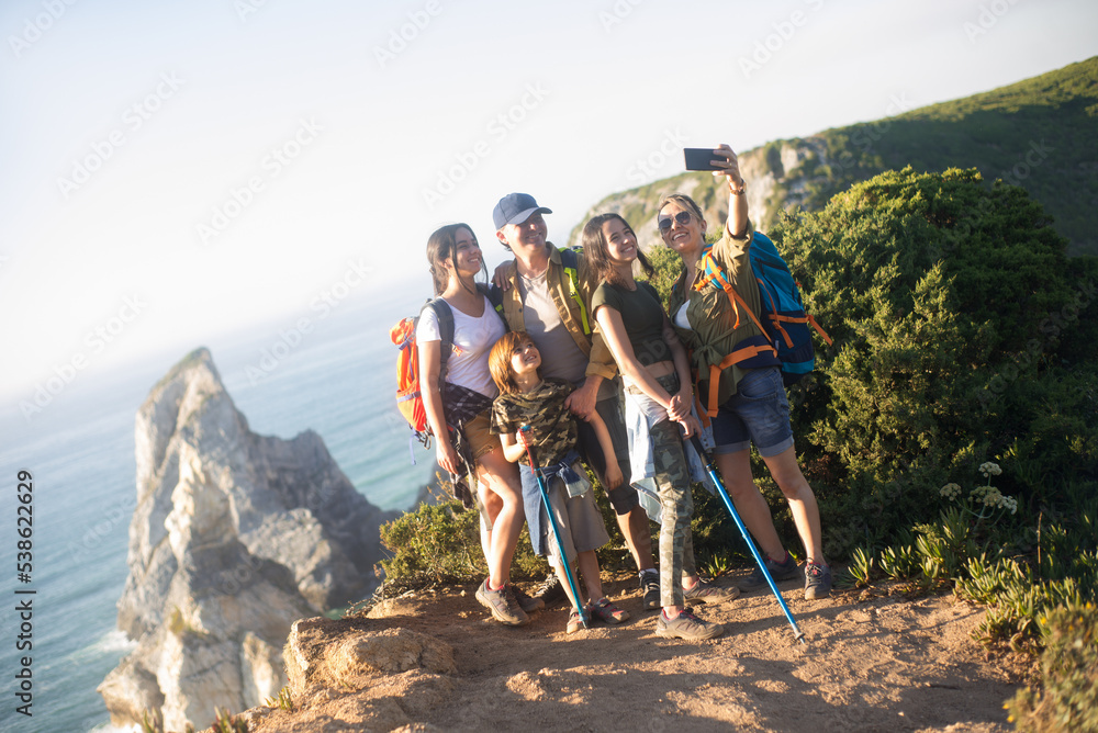 Happy family taking selfie with smartphone outdoor. Portrait of parents hiking with daughters and son in mountains in summer. Active family weekend concept