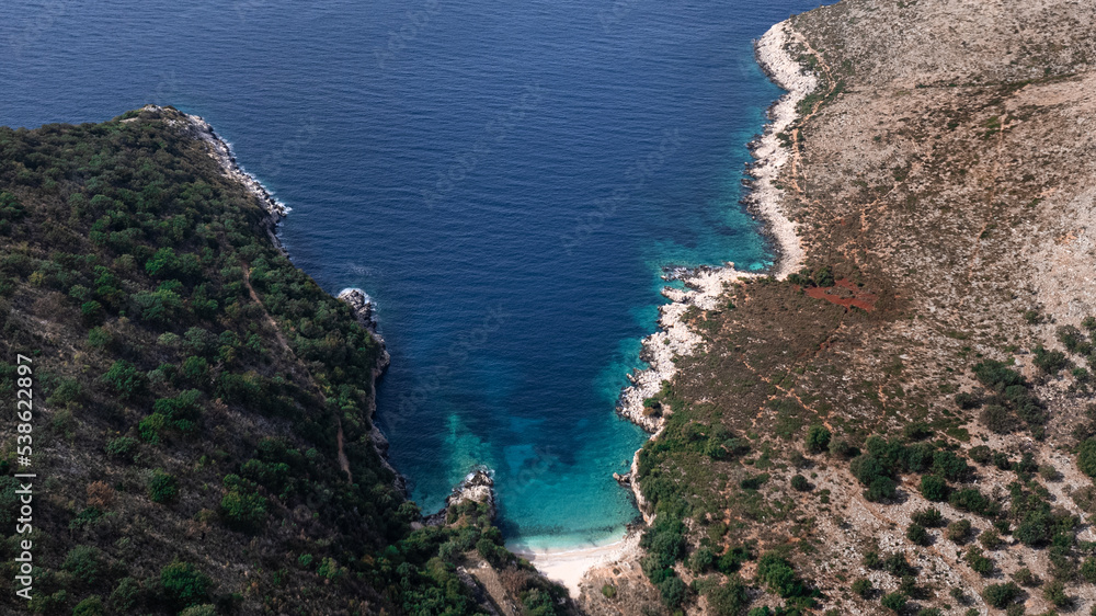 View from above of a bay with blue and turquoise water and a beach, surrounded by hills in Albania