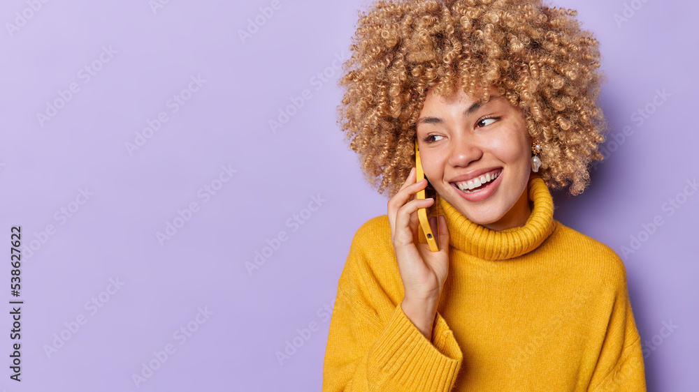 Horizontal shot of happy curly haired woman has telephone conversation talks on mobile phone answers call looks gladfully wears yellow jumper isolated over purple background empty space for your promo Horizontal shot of happy curly haired woman has telephone conversation talks on mobile phone answers call looks gladfully wears yellow jumper isolated over purple background empty space for your promo
