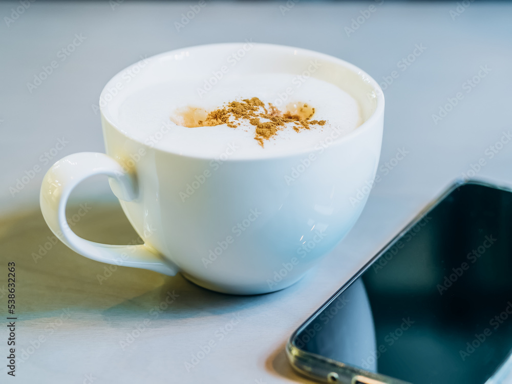 A cup of latte with cinnamon on the table near the smartphone. Selective focus, close-up A cup of latte with cinnamon on the table near the smartphone. Selective focus, close-up