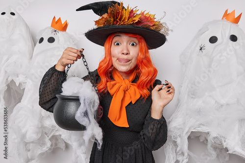 Happy holiday concept. Lovely Asian woman licks lips holds caulderon wears witch hat black dress and orange bow around neck poses against scary decor prepapares for Halloween frightens during night photo