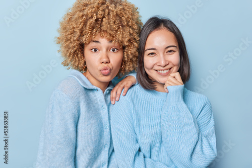 Portrait of diverse women pose together for making photo smile pleasantly keep lips folded dressed in casual sweaters isolated over blue background. Two female friends have friendly relationships