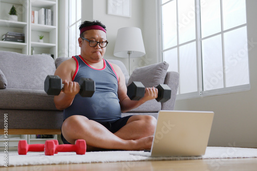 Fat middle-aged asian man in sportswear using dumbbell exercising following trainer online via computer laptop at home photo