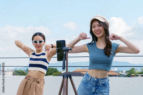 Young Asian woman with her friend tiktoker created her dancing video by smartphone camera together on rooftop outdoor at sunset. To share video to social media application photo