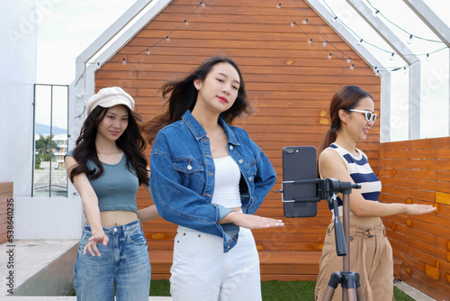 Young Asian woman with her friend tiktoker created her dancing video by smartphone camera together on rooftop outdoor at sunset. To share video to social media application photo