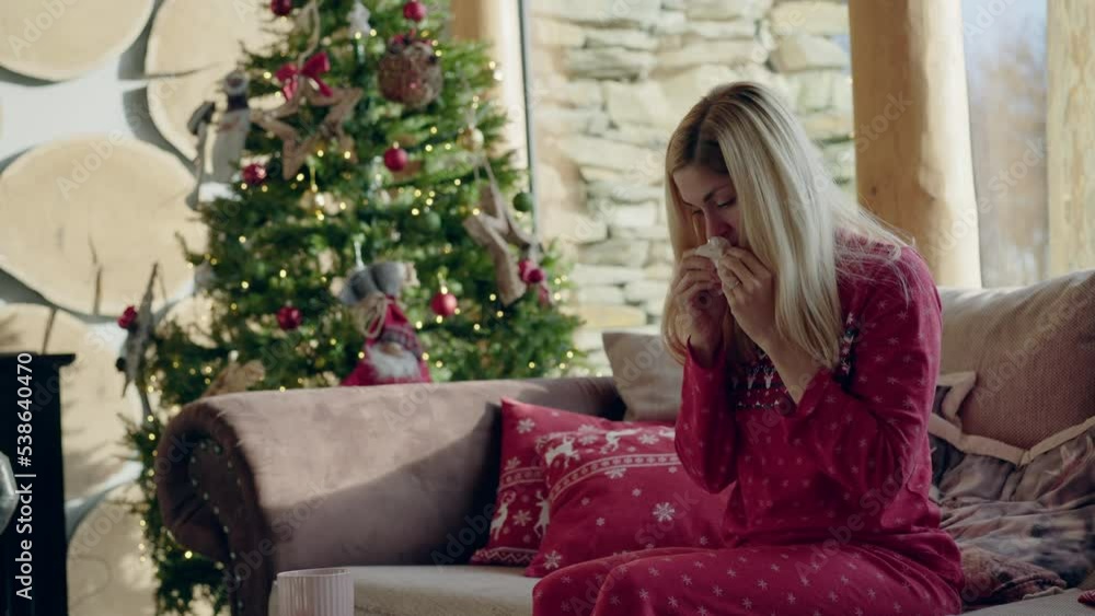 A blonde woman sitting on a sofa having a cold and wiping her nose with a Christmas tree in the background at daylight A blonde woman sitting on a sofa having a cold and wiping her nose with a Christmas tree in the background at daylight