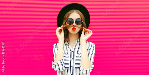 Portrait of beautiful young woman showing mustache her hair blowing red lips sending sweet air kiss wearing black round hat, sunglasses on pink background
