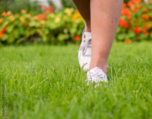 women's feet in white sneakers on the grass on the lawn against the background of blurred flowers