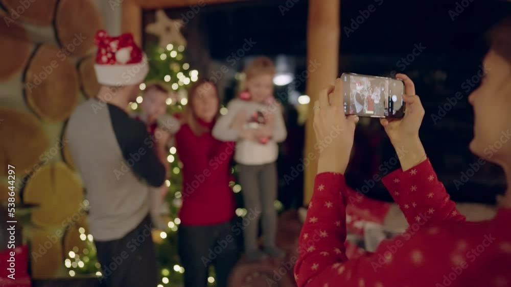 Bokeh - Woman taking photos of a young family of four with her smartphone in front of a Christmas tree in a beautiful cottage Bokeh - Woman taking photos of a young family of four with her smartphone in front of a Christmas tree in a beautiful cottage